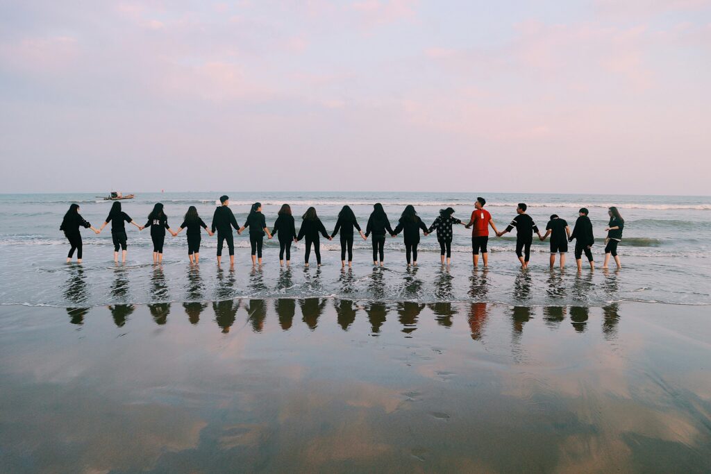 A large group holding hands at the beach during sunset, reflecting in the water.